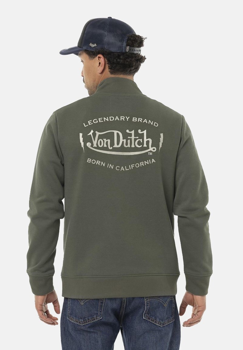 Homme portant une veste olive de la marque Von Dutch avec un logo et le texte "Legendary Brand Born in California" au dos, un jean sombre et une casquette marine.