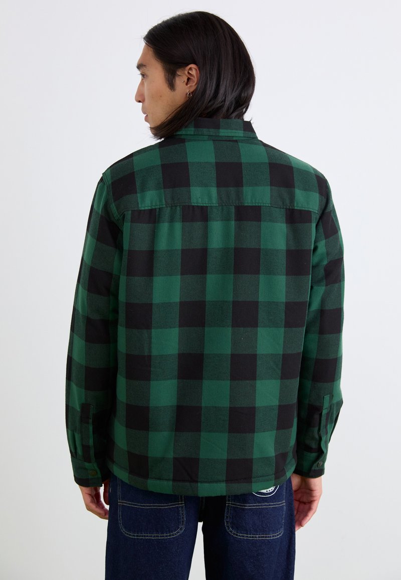 Camicia in flanella a quadri verdi e neri con colletto bottone, maniche lunghe e orlo leggermente curvo, realizzata in tessuto morbido.
