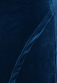 Fluweel stof in diepblauw met een gladde textuur. Heeft een subtiele glans en een naad detail langs de rand, wat het ontwerp versterkt.