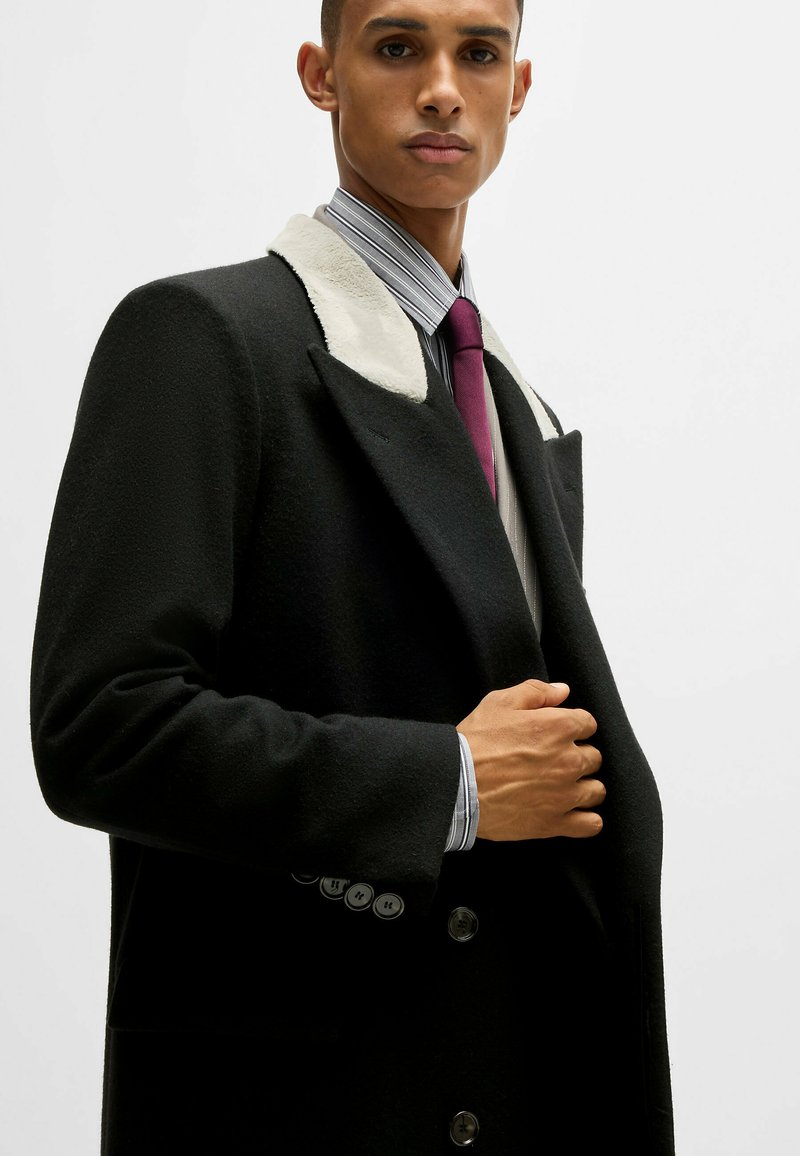 Schwarzer Mantel mit einem weißen Lammfellkragen, doppelreihigem Design und glänzenden Knöpfen. Das Model trägt ein gestreiftes Hemd und eine kastanienbraune Krawatte.