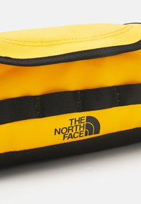 Κίτρινη νάιλον τσάντα με μαύρες λεπτομέρειες, διαθέτοντας ραμμένο άκρο και λογότυπο εκτυπωμένο σε μαύρο. Περιλαμβάνει ιμάντα για μεταφορά.