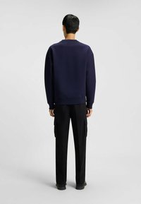 En man står med ryggen mot kameran iklädd en mörk marinblå sweatshirt och svarta cargobyxor med sidofickor mot en enfärgad bakgrund.