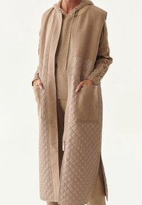 Beige dlouhý kabát s kapucí, prošívaná textura na spodní polovině, dvě přední kapsy a zapínání na zip. Nositý přes odpovídající outfit.
