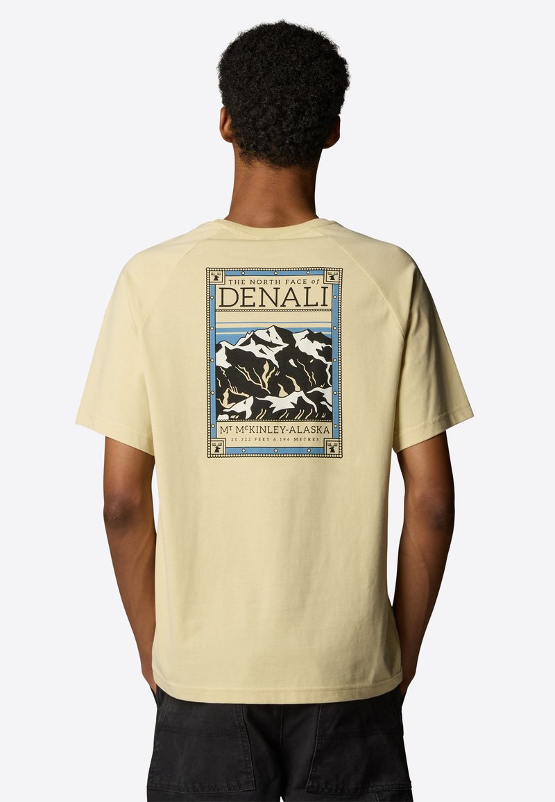 Hombre que lleva una camiseta amarilla claro con un gráfico de montañas nevadas y el texto "La cara norte de Denali, Mt McKinley, Alaska" en la espalda.