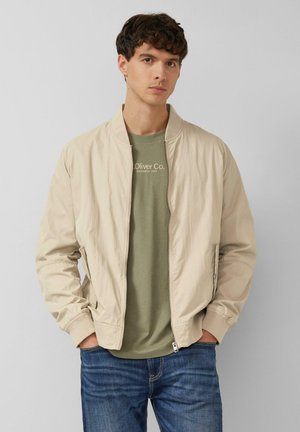 Junger Mann, der eine beige Bomberjacke über einem olivgrünen Oliver Co. T-Shirt und blauen Jeans trägt, mit den Händen in den Taschen stehend.