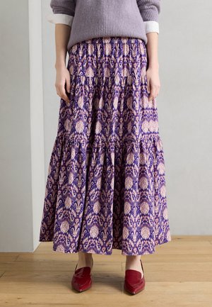 Kobieta w długiej spódnicy w fioletowe wzory, liliowym swetrze z podwiniętymi białymi rękawami i czerwonych butach z zaostrzonym noskiem stoi na drewnianej podłodze.