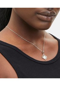 Zilveren hangerketting met een rechthoekige charm met een geometrisch ontwerp, hangend aan een delicate ketting tegen een zwarte top.