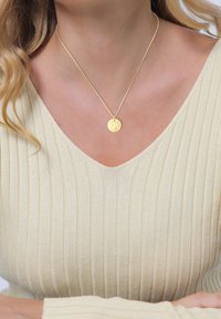 Zlatý prívesok na jemnom reťazci, so štruktúrovaným kruhovým príveskom. Nosený s bledým rebrovaným topom s výstrihom do "v".