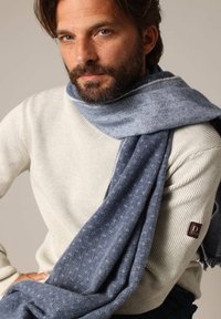 Pull en laine côtelé gris clair avec texture, associé à une écharpe bleu doux arborant un motif subtil. Jean foncé et patch de marque sur le pull.