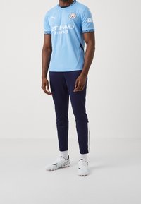 Vaaleansininen Manchester Cityn pelipaita mustilla yksityiskohdilla ja logolla, yhdistettynä tummansinisiin urheiluhousuihin, joissa on valkoiset sivuraidat, ja valkoisiin jalkapallokenkiin.