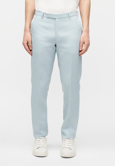 Άνδρας με ανοιχτό μπλε παντελόνι στενής εφαρμογής και λευκά αθλητικά παπούτσια, στέκεται μπροστά σε απλό λευκό φόντο.