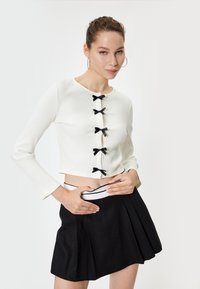 Top corto blanco de canalé con mangas largas, que presenta acentos de lazo negro en el centro. Combinado con una falda plisada negra y una cinturilla blanca.