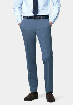 Mann trägt maßgeschneiderte blaue Anzughose, weißes Hemd, marineblaue Krawatte, schwarzen Gürtel und braune Wildleder-Loafer, steht mit den Händen in den Taschen.