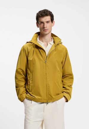 Mann trägt eine senfgelbe Jacke mit Reißverschluss und Kragen über einem weißen Hemd und hellen Hosen, Hände in den Taschen, schaut nach vorne.
