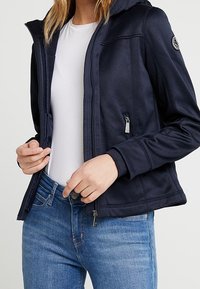 Marinblå hooded jacka i slätt, elastiskt material med fickor med dragkedja och figurnära passform, bärs över en vit skjorta och blå jeans.