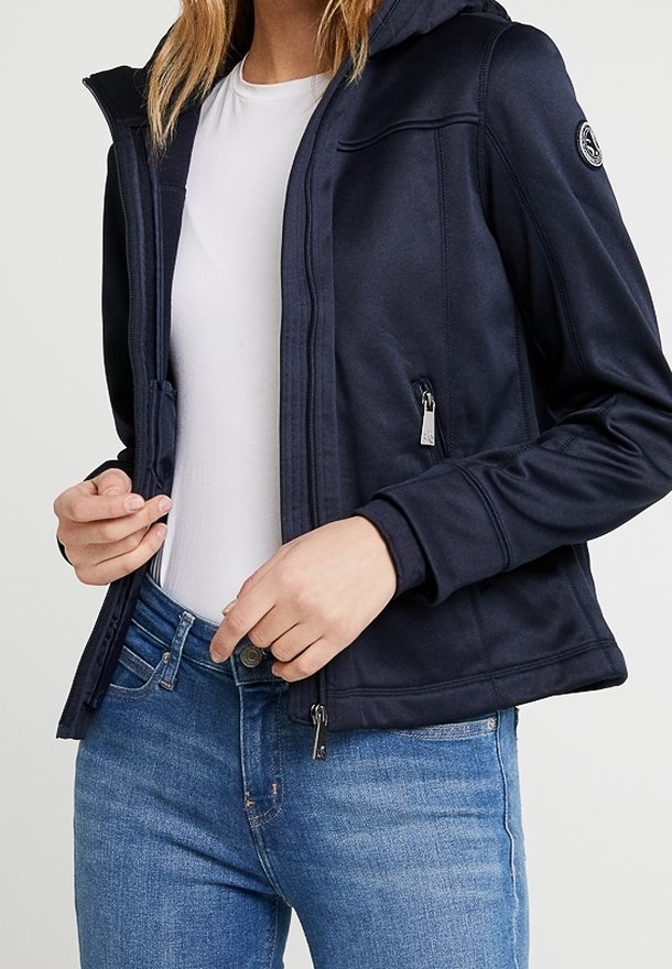 Marinblå hooded jacka i slätt, elastiskt material med fickor med dragkedja och figurnära passform, bärs över en vit skjorta och blå jeans.