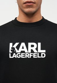 Κοντινό πλάνο ενός ατόμου που φοράει μαύρο πουκάμισο με το λευκό κείμενο "KARL LAGERFELD" και ένα σιλουέτα λογότυπο στην αριστερή πλευρά.