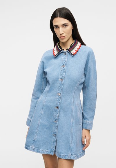 Γυναίκα που φοράει ένα ελαφρύ φόρεμα από τζιν μπλε χρώμα με κουμπιά μπροστά και πολύχρωμο, με μοτίβο γιακά.