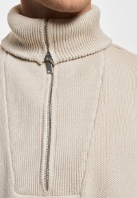 Gros plan sur un pull en tricot beige avec un col côtelé haut et une fermeture éclair argentée partiellement zippée à l'avant.