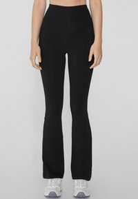 Pantalons de yoga noirs évasés avec une taille haute, texture lisse et une couture en forme de V à l'avant, accompagnés de chaussures de sport blanches.