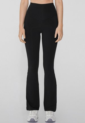 Zwarte flared yoga pants met een hoge tailleband, een gladde textuur en een V-vormige naad aan de voorkant, gecombineerd met witte sportschoenen.