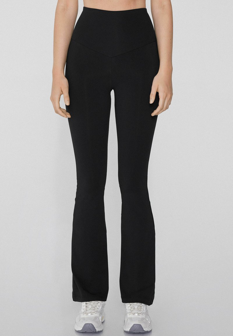 Pantalons de yoga noirs évasés avec une taille haute, texture lisse et une couture en forme de V à l'avant, accompagnés de chaussures de sport blanches.