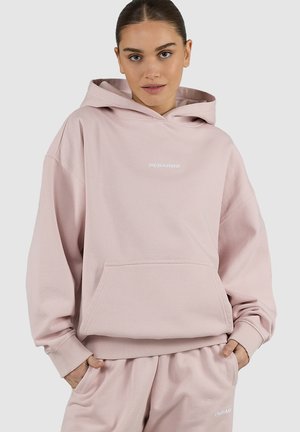 Ung kvinde iført lyserød hættetrøje og matchende joggingbukser med hænderne i lommerne, stående mod en ensartet lys baggrund.