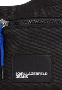 Black textured fabric bag with silver zipper and blue cord accent. Features a rectangular black label with "KARL LAGERFELD JEANS" in white text.