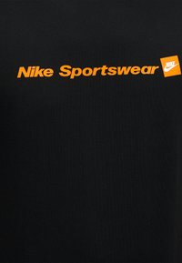 Μαύρο ύφασμα με πορτοκαλί κείμενο "Nike Sportswear" και πορτοκαλί τετράγωνο λογότυπο με λευκό σύμβολο Nike swoosh και κείμενο.