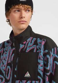 Veste en polaire avec un motif tourbillon coloré en bleu et rose, accents noirs unis, col haut et une poche poitrine zippée.