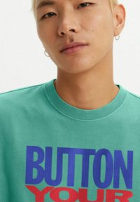 Turquoise cotton T-shirt features bold text "BUTTON YOUR" in blue and "YOU" in red. Round neckline and short sleeves.