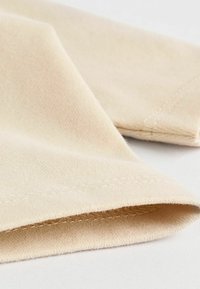 Beige Stoff mit einer weichen Textur, der einen gefalteten Saum mit sichtbaren Nähten aufweist. Eine Nahaufnahme betont die glatte Oberfläche und die klaren Kanten.