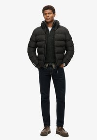 Sort pufferjakke med høj krave, struktureret striktrøje under, mørke jeans og brune snørebåndssko. Modellen står oprejst.