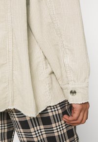 Camisa de pana color beige con rayas verticales, dobladillo deshilachado y detalles de botones a presión plateados, combinada con pantalones de cuadros negros y marrones.