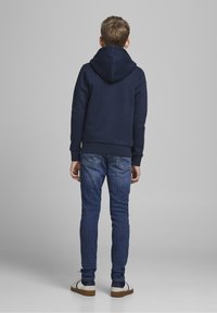 Marineblaues Hoodie mit großer Kapuze, getragen mit schmal geschnittenen blauen Jeans und weißen Sneakers mit dunklen Akzenten und einer sandfarbigen Sohle.