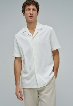 Uomo che indossa una camicia bianca a maniche corte con bottoni e texture e pantaloni beige, in piedi davanti a uno sfondo grigio chiaro e uniforme.