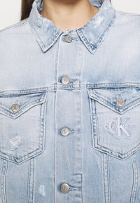 Giacca di jeans azzurro chiaro con colletto classico, dettagli vissuti, bottoni argentati e logo ricamato Calvin Klein sulla tasca del petto.
