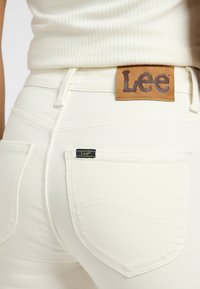 Jeans de mezclilla blanca con un acabado texturizado. Las características incluyen una etiqueta de cuero marrón en la cintura y un pequeño logotipo negro en el bolsillo trasero.