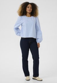 Blusa azzurra chiara con maniche a sbuffo, orlo scalloppato e rifiniture bianche, abbinata a jeans in denim scuro e scarpe chiare.