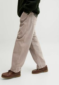 Beżowe spodnie z szerokimi nogawkami wykonane z bawełny, z widocznymi szwami i tylną kieszonką, połączone z brązowymi butami na sznurówki. Gładka tekstura.