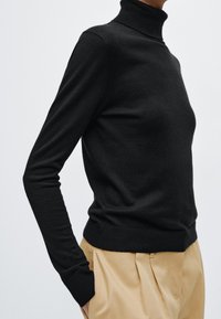 Czarny sweter z golfem wykonany z miękkiego materiału, o dopasowanym kroju i ściągaczowych mankietach. W komplecie z jasnobeżowymi spodniami.
