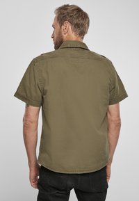 Cămașă cu mânecă scurtă de culoare verde măslin, cu guler cu nasturi, cusături vizibile și un material neted, ușor. Vederea din spate arată un design simplu.