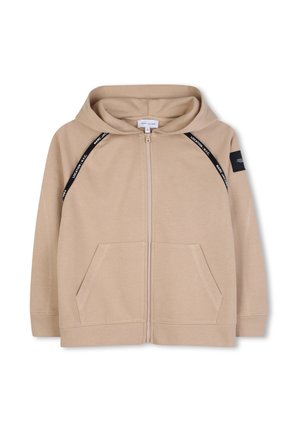Sweat à capuche beige zippé avec poches avant, bordure diagonale noire avec texte blanc, poignets côtelés et un patch noir sur la manche gauche.