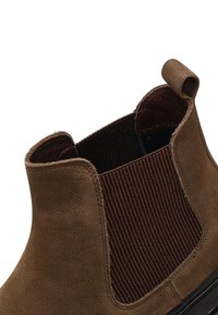 Stivale alla caviglia in camoscio marrone con pannello laterale a costine, inserto elastico e punta arrotondata. Texture liscia con una linguetta per una facile calzata.