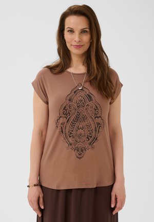 Frau mit langen braunen Haaren, die ein braunes kurzärmeliges Shirt mit einem kunstvollen schwarzen Paisley-Muster und einer silbernen Halskette trägt.