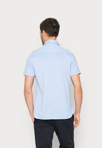 Uomo con capelli ricci scuri, indossa una camicia a maniche corte azzurro chiaro e pantaloni neri, in piedi con le spalle rivolte verso uno sfondo chiaro e semplice.