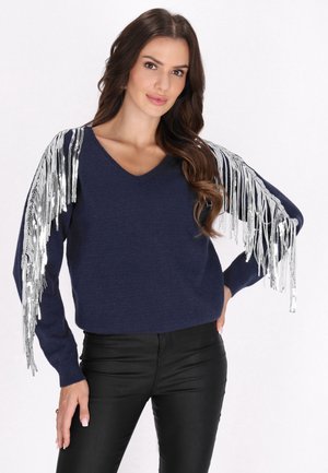 Jonge vrouw met lang bruin haar draagt een marineblauwe trui met zilveren franjes op de mouwen en een zwarte leren broek, staand tegen een witte achtergrond.