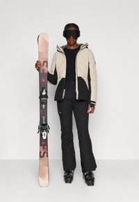 O equipamento de ski inclui uma jaqueta isolada em bege e preto, calças pretas e botas de ski, com uma prancha de snowboard na mão mostrando detalhes gráficos.