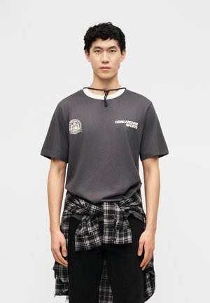 Νεαρός άνδρας που φοράει σκούρο γκρι μπλουζάκι με την επιγραφή "CORE ARCHIVE SPORTS" και ένα ασπρόμαυρο καρό πουκάμισο δεμένο γύρω από τη μέση του, με λευκό φόντο.