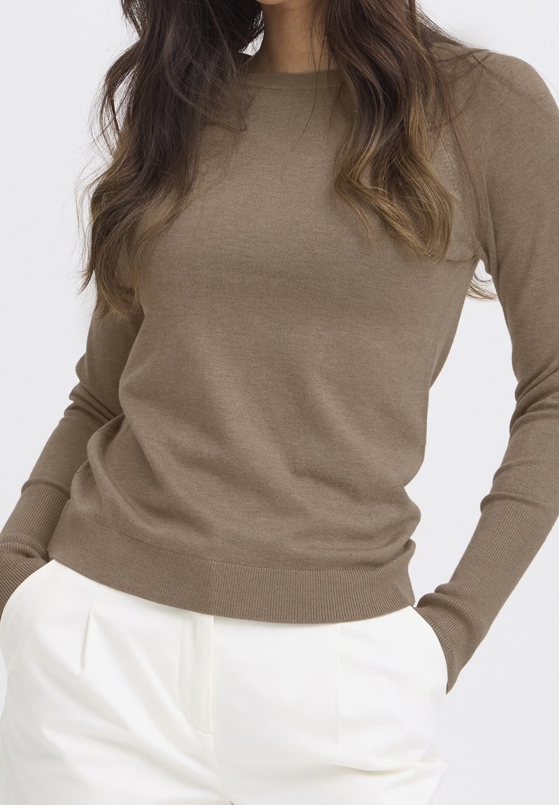 Femme aux longs cheveux bruns portant un pull moulant marron clair et un pantalon blanc, les mains dans les poches, sur un fond uni.